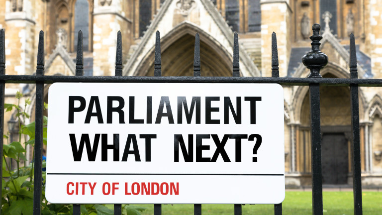 Sign in the style of a London Street sign asking Parliament, what next? Black white and red banner against wrought iron railings, with Westminster Abbey in the background.